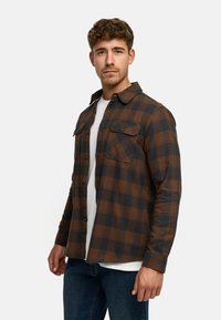 Brązowa i czarna koszula flanelowa w kratkę z kołnierzykiem zapinanym na guziki, podwójnymi kieszeniami na piersi i długimi rękawami, noszona na białym T-shircie.