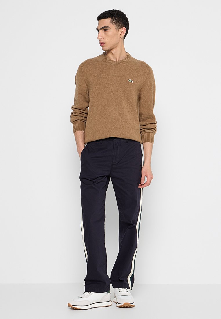 Maglione marrone lavorato a maglia con polsini a coste, abbinato a pantaloni blu navy con strisce laterali bianche e verdi. Modello che indossa sneakers bianche.