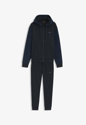 Sweat à capuche zippé bleu marine avec manches texturées et pantalon de jogging assorti, tous deux ornés de petits logos « BOSS » sur la poitrine et la cuisse.