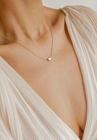 Goldene Halskette mit einem kleinen Herzanhänger an einer zarten Kette, die an einem nackten Hals getragen wird. Weißes, plissiertes Kleidungsstück im Hintergrund.