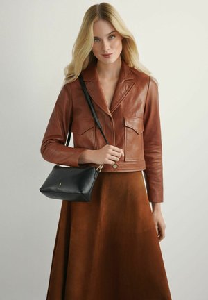 Blondynka ubrana w brązową skórzaną kurtkę i spódnicę, trzymająca czarną torebkę na ramię z złotym zapięciem.