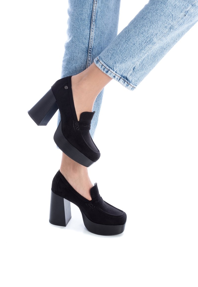 Femme portant un jean bleu clair et des mocassins noirs à talon compensé, pied levé sur fond blanc.