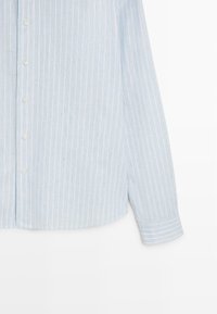 Chemise bleu clair à manches longues avec rayures blanches verticales, boutons sur le devant et poignets boutonnés, présentée sur un fond blanc.