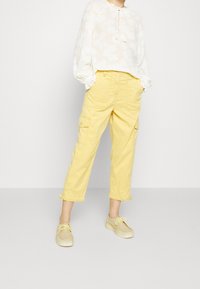 Vrouw in een losse witte blouse met patroon, gele cropped cargobroek en beige veterschoenen, staand met handen in de zakken.
