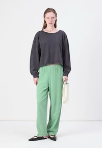 Szürke cropped pulcsi, zöld textúrájú sportnadrág és fekete lapos cipők. A modell egy kis bézs táska mellett áll. Egyszerű, laza öltözet.