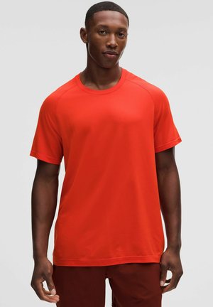Junger Mann mit dunkler Haut, der ein leuchtend oranges kurzärmeliges Sportshirt und dunkelrote Shorts trägt, steht vor einem einfarbigen hellen Hintergrund.
