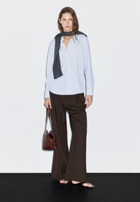 Camicia a maniche lunghe azzurra con scollo a V, abbinata a pantaloni larghi marrone scuro. Una sciarpa grigia scura è drappeggiata attorno al collo, mentre si tiene una borsa a tracolla marrone.