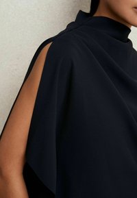 Detailní záběr na modelku, která má na sobě tmavý, elegantní oděv s vysokým výstřihem a odkrytým ramenem, předvádějící texturovanou látku na jemném pozadí.