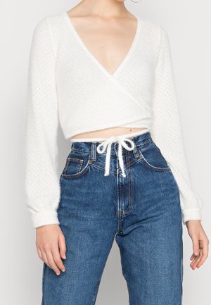Mujer con top corto de punto borroso blanco con escote en V y mangas largas, y vaqueros azules de talle alto atados con un lazo de cuerda blanca.