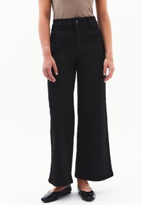 Vaqueros negros de pierna ancha, hechos de denin resistente, con una cintura alta, bolsillos frontales y una textura suave, combinados con zapatos planos negros.