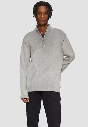 Grijze pullover met een hoge hals en een halve rits aan de voorkant. Geribbelde textuur en lange mouwen. Gepaard met donkere cargobroeken.