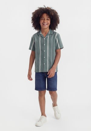 Bambino sorridente con capelli ricci che indossa una camicia verde a righe a maniche corte con bottoni, pantaloncini di jeans e scarpe da ginnastica bianche, che cammina in avanti.