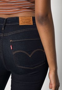 Jeans de mezclilla azul oscuro con una etiqueta de parche de cuero, costuras doradas y un diseño distintivo en el bolsillo trasero que presenta una costura curva.