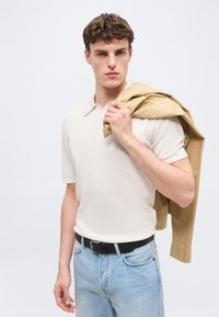 Jeune homme portant une chemise blanche à manches courtes et un jean bleu clair, tenant une veste beige sur son épaule, devant un fond uni.