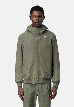 Uomo che indossa una giacca verde con cappuccio e pantaloni verdi coordinati, in piedi dritto contro uno sfondo grigio chiaro uniforme.