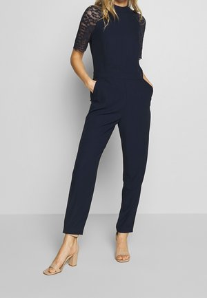 Marineblauer Jumpsuit mit kurzen Spitzenärmeln, tailliertem Oberteil und schmalen Beinen. Mit seitlichen Taschen und einem Rundhalsausschnitt. Beige Blockabsatzsandalen.