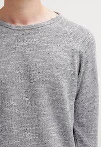 Zbliżenie osoby w jasnoszarym, dzianym swetrze z okrągłym dekoltem na jednolitym białym tle.