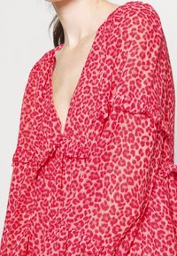 Donna che indossa un vestito leopardato rosa e rosso con dettagli arricciati e scollatura a V profonda, su uno sfondo bianco semplice.