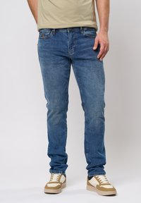 Jeans de mezclilla azul para hombre con corte recto, cinco bolsillos y un acabado desgastado. Combinados con zapatillas beige y blancas.