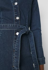 Jeansjacka med en mörkblå tvätt, prydd med silverknappar, bälte i midjan och uppvikta ärmar med kontrasterande stygn.