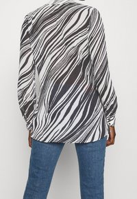 Persona che indossa una blusa trasparente a righe zebrate in bianco e nero con maniche lunghe e jeans blu, vista da dietro su uno sfondo bianco semplice.