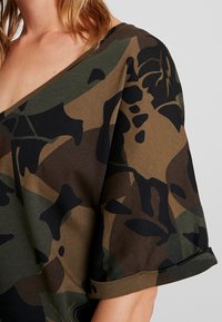 Topp med camouflagemönster och V-ringning, korta ärmar och uppvikta ärmslut; färgerna inkluderar grön, brun och svart i varierande former.