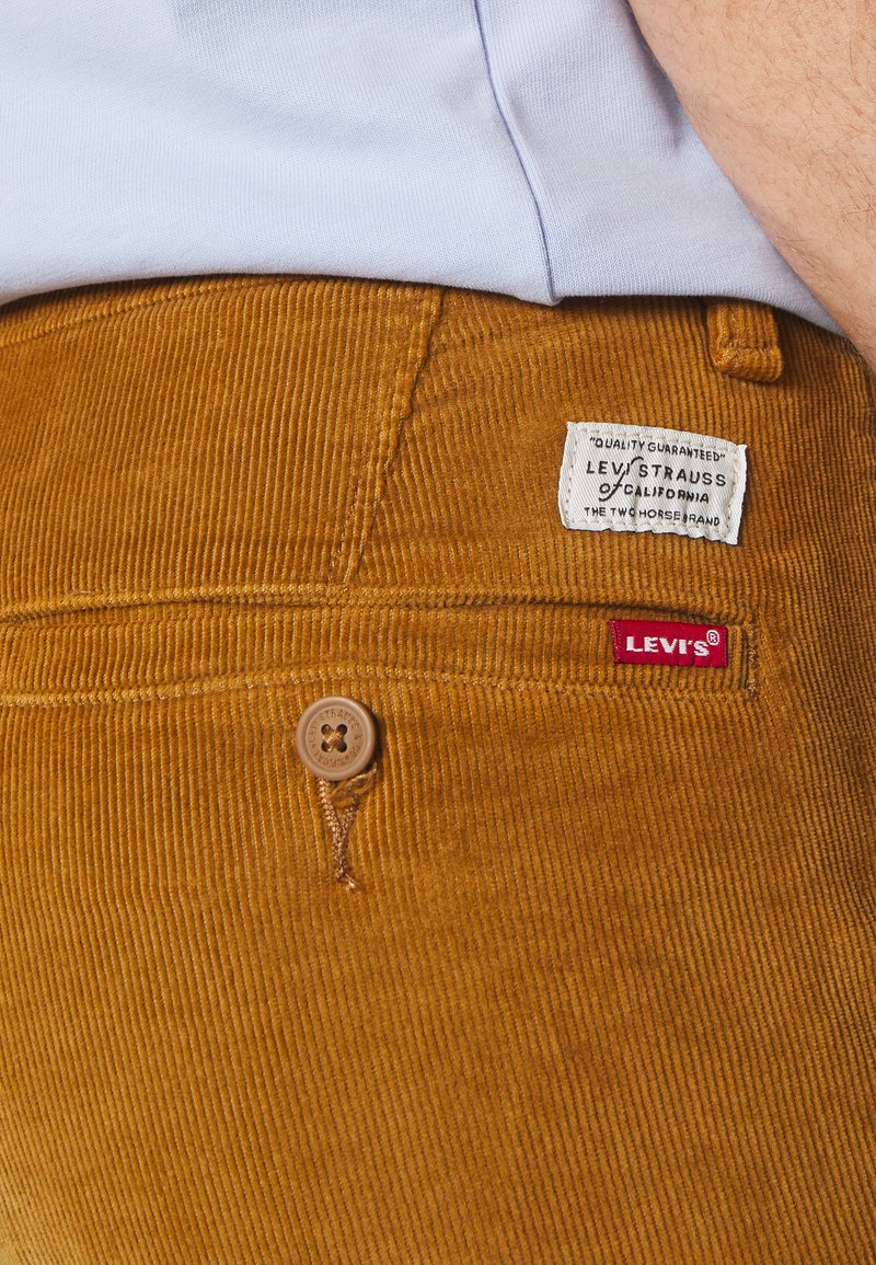Pantalons en velours côtelé marron avec une texture à côtes horizontales, une fermeture à bouton et des étiquettes de marque : une étiquette blanche avec "Levi Strauss" et une étiquette rouge "Levi's".