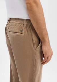 Brązowe spodnie sztruksowe z pionowymi prążkami, elastycznym pasem, bocznymi kieszeniami oraz małym metalowym logo na tylnej kieszeni.