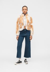 Γυναίκα με πλεξούδες που φοράει μπεζ φλις μπουφάν με σχέδια, λευκό κοντό τοπ, τζιν παντελόνι ευρέος ποδιού σε μπλε χρώμα, λευκές κάλτσες και μαύρα αθλητικά παπούτσια, όρθια.