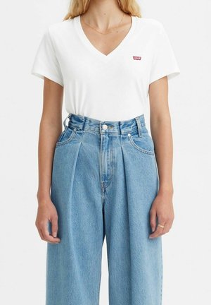 Woman wearing white V-neck Levi’s T-shirt tucked into high-waisted, pleated, light blue denim jeans, with a thin gold necklace and ring.