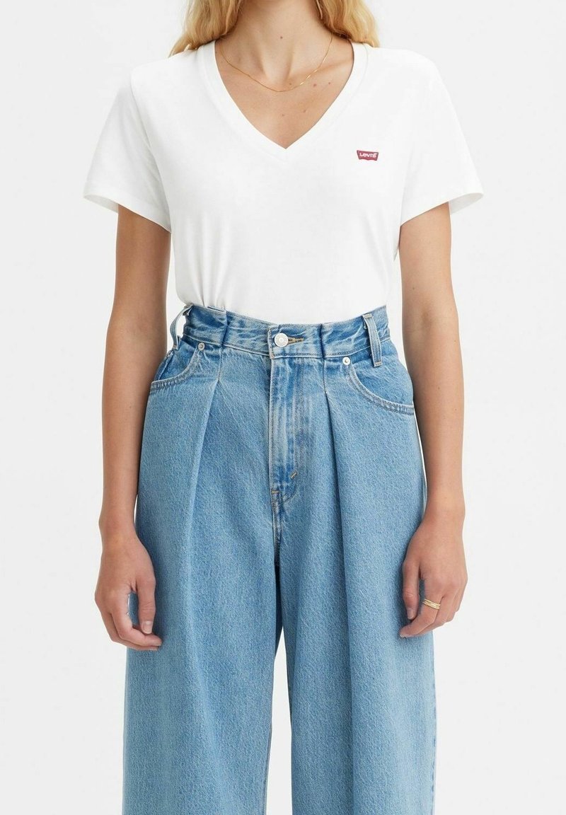Femme portant un t-shirt Levi’s blanc à col en V rentré dans un jean taille haute, plissé, en denim bleu clair, avec un fin collier en or et une bague.