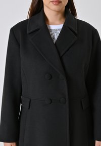 Cappotto nero doppiopetto con grandi bottoni, colletto a rever e spalle strutturate. Realizzato in tessuto testurizzato con una vestibilità sartoriale.