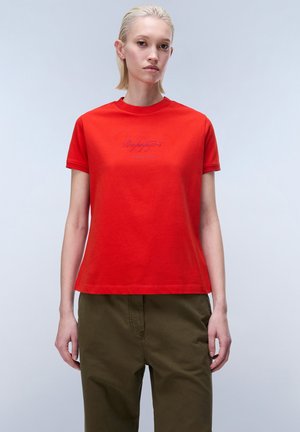 Blonde woman wearing a red short-sleeve T-shirt with subtle text and olive-green pants, standing against a light gray background.