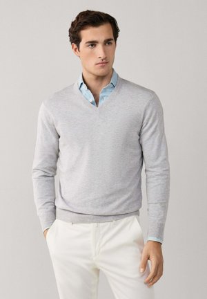 Junger Mann, der einen hellgrauen V-Ausschnitt-Pullover über einem blauen Hemd und weißen Hosen trägt, steht mit einer Hand in der Tasche.