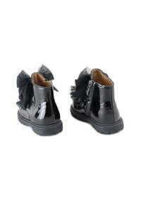 Botines de charol negro con un lazo de tul negro mate en la parte trasera, cierre lateral con cremallera y suela de goma texturizada.