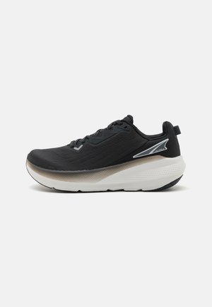 Chaussure de sport noire avec une tige texturée, semelle blanche et beige, bout rond et accents minimalistes comprenant un détail réfléchissant sur le côté.