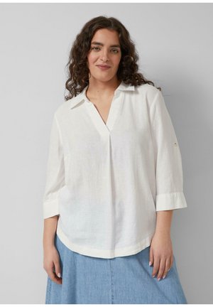 Kobieta z kręconymi ciemnymi włosami, ubrana w luźną białą bluzkę z rękawami do łokcia oraz jasnoniebieską spódnicę jeansową, stojąca na jednolitym tle.