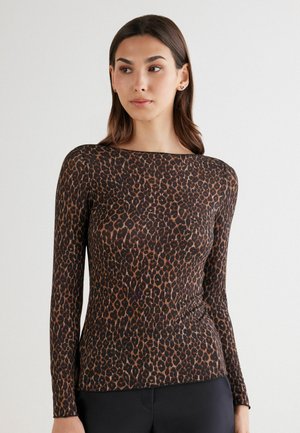 Vrouw in een nauwsluitend, lang mouw leopardprint shirt en zwarte broek, staand tegen een effen lichte achtergrond.