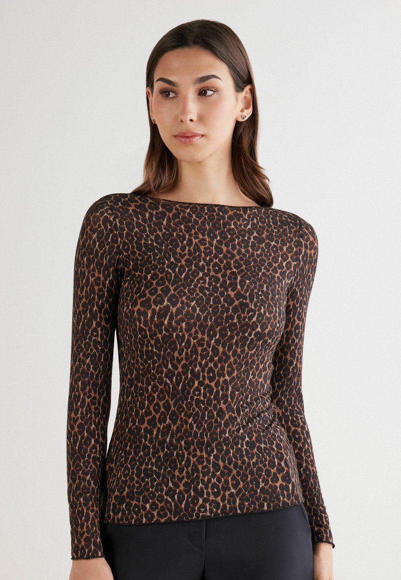 Vrouw in een nauwsluitend, lang mouw leopardprint shirt en zwarte broek, staand tegen een effen lichte achtergrond.
