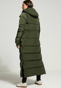 Giacca a vento imbottita lunga di colore verde oliva con cappuccio, presenta cuciture a zigzag e fessure laterali per la mobilità, abbinata a scarpe leggere.