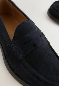 Mocasines de ante navy con un borde superior cosido, acentos texturizados y un forro de cuero suave. La suela es marrón oscuro con un ligero brillo.