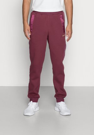 Homme portant un jogging bordeaux avec des poches latérales bordeaux brillantes, logo Adidas et des baskets blanches, debout devant un fond uni.