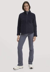 Marineblauwe fleece zip-up jas met hoge kraag, voorzien van twee voorzakken, gecombineerd met slim-fit grijze broek en lichtgekleurde sportschoenen.