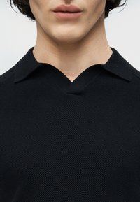 Κοντινό πλάνο ενός ατόμου που φοράει υφασμένο μαύρο πουκάμισο με γιακά, εμφανίζοντας τον λαιμό, τα χείλη και μέρος του πηγουνιού πάνω σε απλό φόντο.