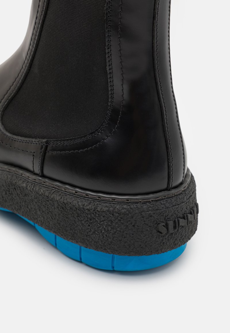 Stivale alla caviglia nero in pelle opaca, con pannelli elasticizzati, suola nera texturizzata e un accent luminoso blu alla base. Dettagli delle cuciture visibili.