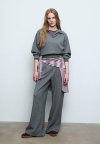 Szary sweatshirt z szerokim kołnierzem i opadającymi ramionami, w połączeniu z szarymi spodniami o szerokich nogawkach. Pasiasty materiał przewiązany w talii dodaje kontrastu.