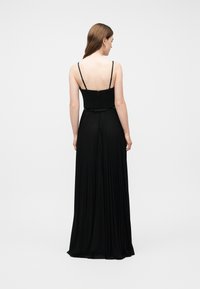 Γυναίκα με μακριά καστανά μαλλιά που φοράει μαύρο αμάνικο πλισέ φόρεμα μάξι, εμφανιζόμενη από πίσω πάνω σε απλό λευκό φόντο.