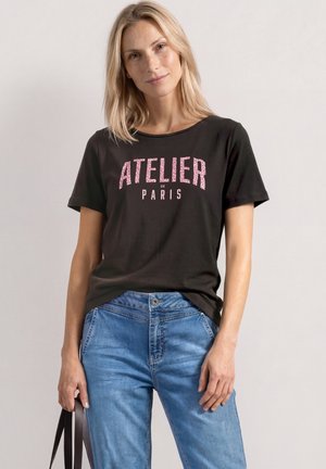 Kvinde med blondt hår iført en sort T-shirt med pink "ATELIER DE PARIS" tekst og blå jeans, der holder en mørk taske-strop i den ene hånd.