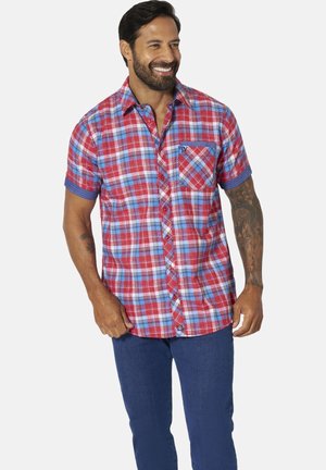 Man smiling wearing a short-sleeve red and blue plaid button-up shirt and blue pants, with tattoos on his right forearm and left arm.
