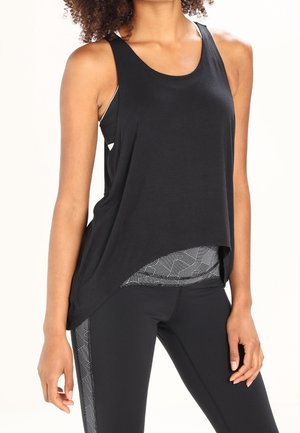 Femme portant un débardeur asymétrique noir sans manches sur des leggings noirs et gris à motifs, debout, une main détendue le long du corps.